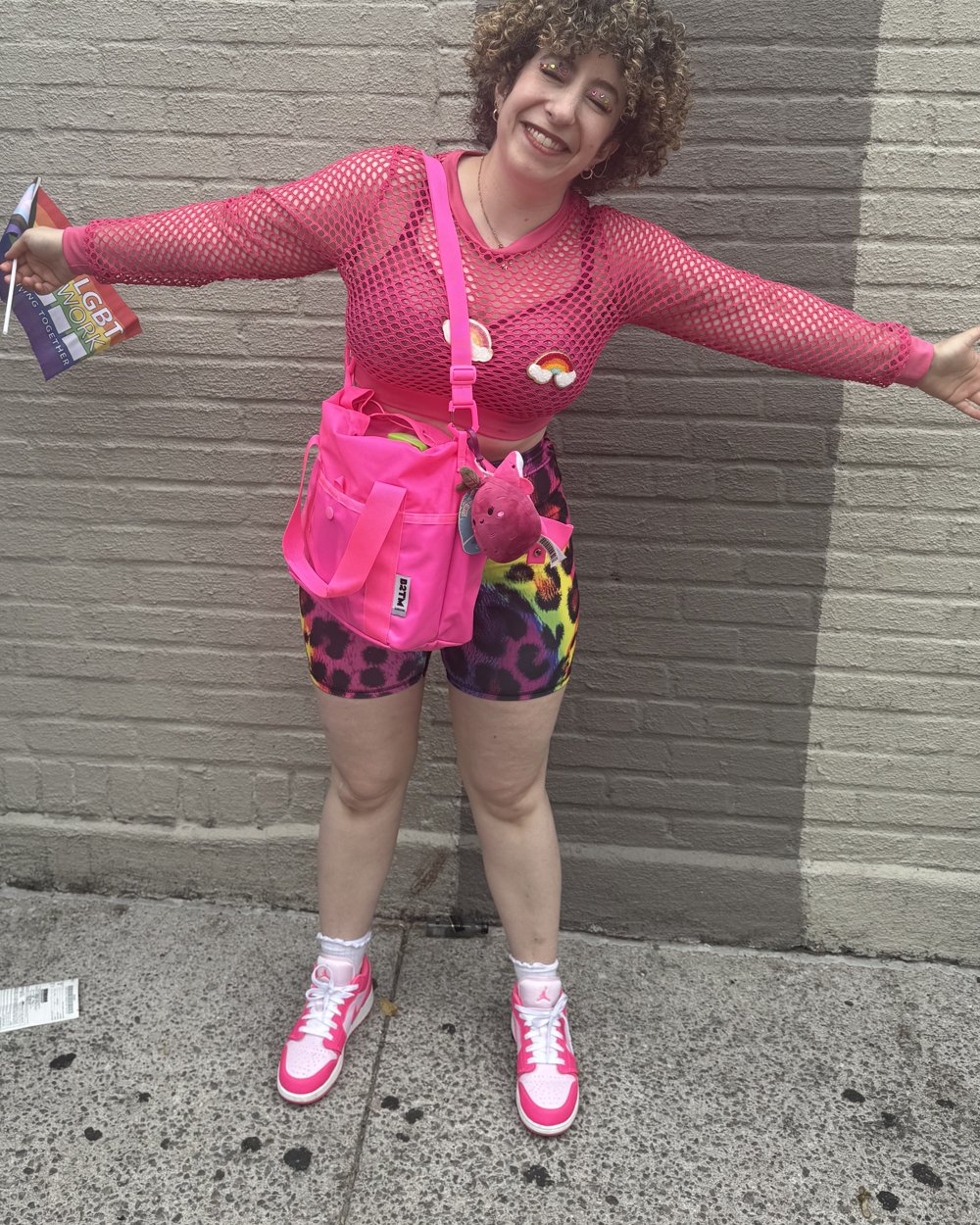 Alex in a hot pink mesh top and rainbow leopard shorts, arms flung wide, holding a pride flag